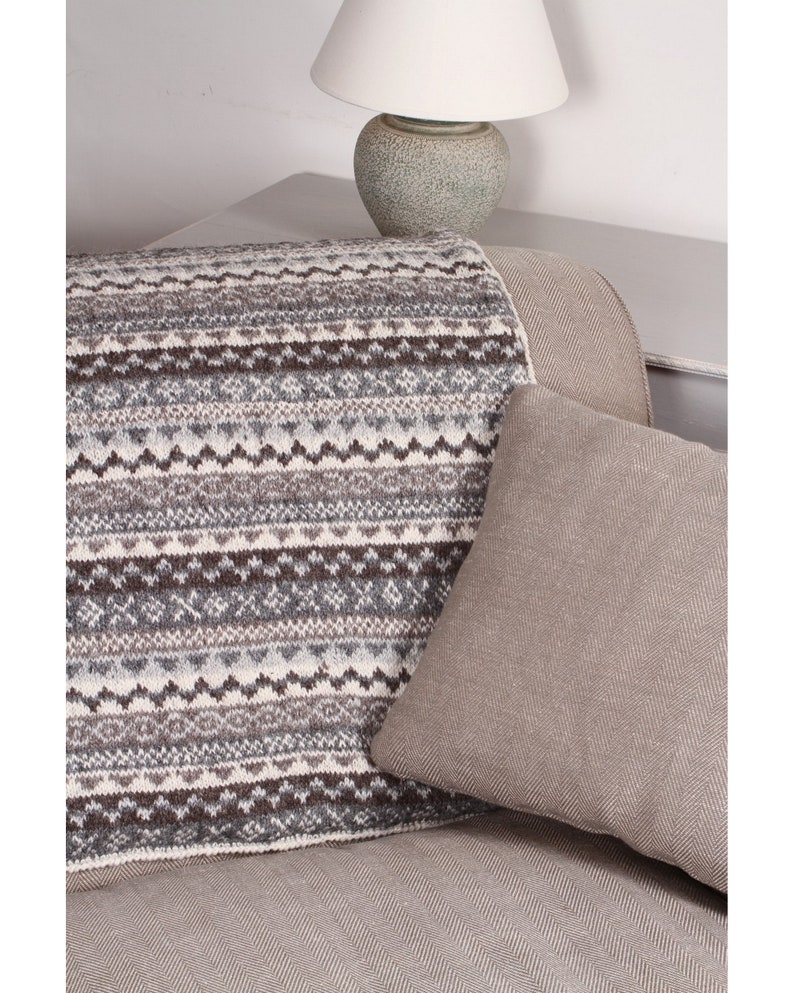 K&ouml;nnte beinhalten: Eine Strickdecke mit geometrischem Muster in Grau-, Braun- und Cremet&ouml;nen liegt &uuml;ber einem Sofa. Ein einfarbiges, hellbraunes Kissen liegt an der Decke. Im Hintergrund befindet sich eine Lampe mit wei&szlig;em Schirm.