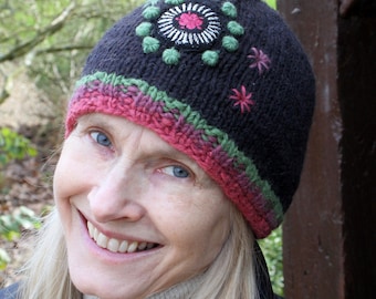 Pode incluir: Um gorro de malha preto com um motivo circular bordado com detalhes em verde e rosa. A borda é decorada com uma faixa horizontal de fio mesclado em rosa e verde, e a lateral inclui dois pequenos detalhes bordados em forma de estrela rosa.