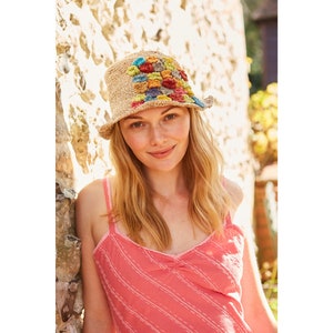 May include: A woman wearing a straw hat with colorful crocheted details. The hat has a wide brim and is decorated with a pattern of squares in various shades of blue, green, yellow, and orange. She is wearing a pink and white striped tank top.