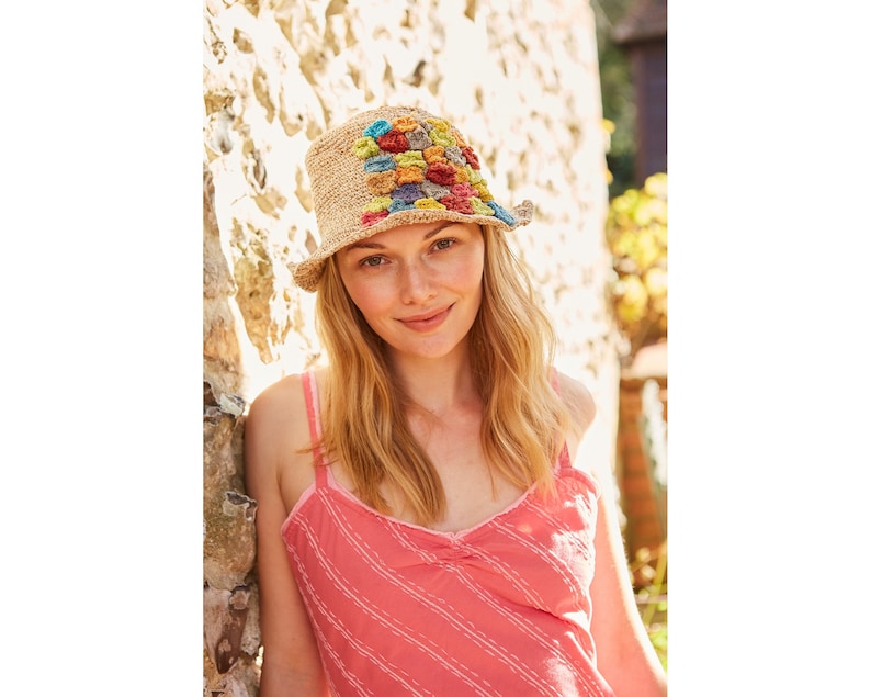May include: A woman wearing a straw hat with colorful crocheted details. The hat has a wide brim and is decorated with a pattern of squares in various shades of blue, green, yellow, and orange. She is wearing a pink and white striped tank top.