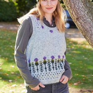 May include: A gray sleeveless sweater with a green and purple thistle pattern. The sweater has a round neckline and is worn over a long-sleeved shirt. The person is wearing a headband with a similar thistle pattern.