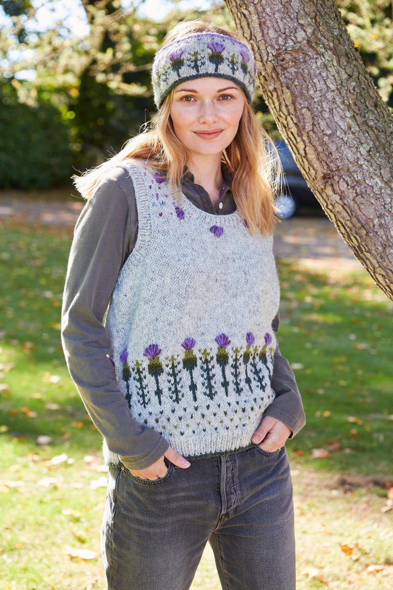 May include: A gray sleeveless sweater with a green and purple thistle pattern. The sweater has a round neckline and is worn over a long-sleeved shirt. The person is wearing a headband with a similar thistle pattern.