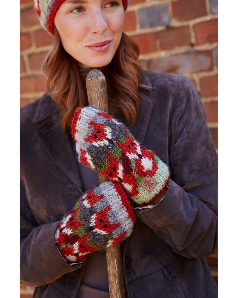 May include: A pair of red, green, and gray knitted mittens with a fox pattern. The mittens have a brown leather tag with a logo on them.