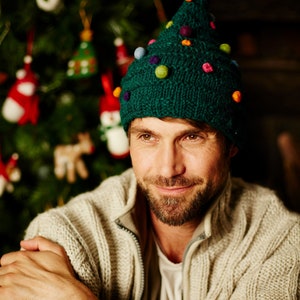 May include: A man wearing a green knitted Christmas tree hat with colorful pom-poms. He is wearing a cream colored sweater and has a beard.
