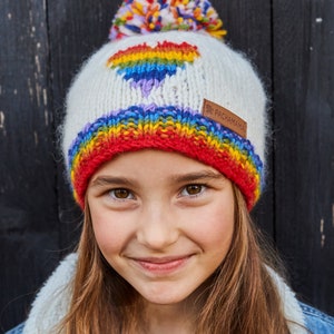 May include: A white knit beanie with a rainbow heart design and a multi-colored pom pom on top. The beanie has a red band at the bottom and the text "Pachamama" on the side.