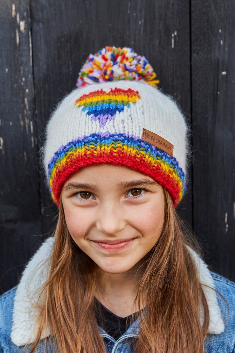 May include: A white knit beanie with a rainbow heart design and a multi-colored pom pom on top. The beanie has a red band at the bottom and the text "Pachamama" on the side.