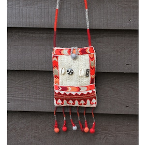 Puede incluir: Una pequeña bolsa blanca tejida con ribetes rojos y naranjas. La bolsa tiene una correa larga y está decorada con pequeñas conchas y cuentas. La bolsa está colgada en una pared de madera.