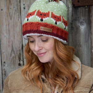 May include: A green and orange knit hat with a pom pom on top. The hat features a repeating pattern of foxes. The hat has a brown leather label that says "Pachamama".