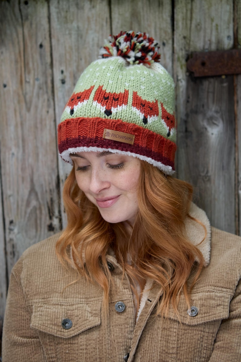 May include: A green and orange knit hat with a pom pom on top. The hat features a repeating pattern of foxes. The hat has a brown leather label that says "Pachamama".