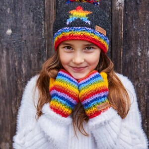 May include: A young person wearing a gray knit hat with a rainbow star and a rainbow striped pom pom. The hat has a brown leather tag with the text "The  Fleece  People". The person is also wearing rainbow striped mittens with a brown leather tag that says "The Fleece People".