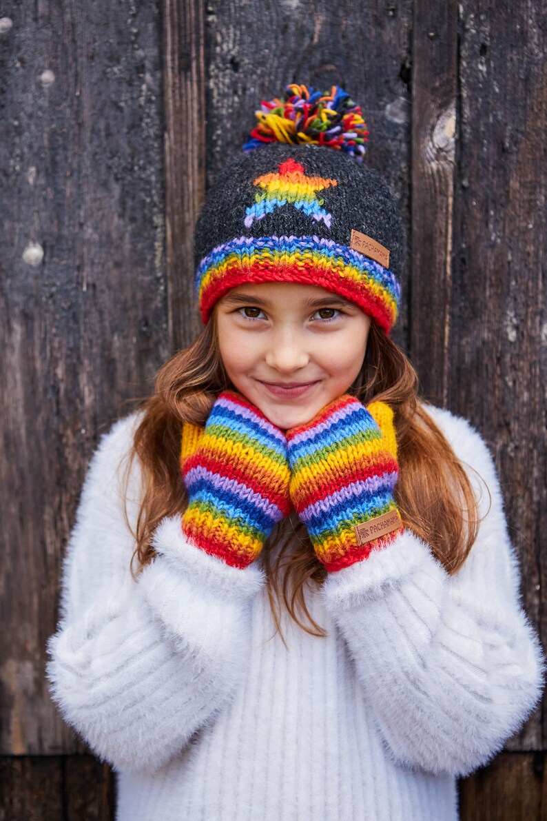 May include: A young person wearing a gray knit hat with a rainbow star and a rainbow striped pom pom. The hat has a brown leather tag with the text "The  Fleece  People". The person is also wearing rainbow striped mittens with a brown leather tag that says "The Fleece People".