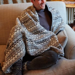 K&ouml;nnte beinhalten: Eine Person sitzt auf einem grauen Sofa und ist in eine Strickdecke mit grauem und wei&szlig;em Muster geh&uuml;llt. Die Person tr&auml;gt einen grauen Rollkragenpullover und eine dunkelgraue Hose. Passende Stricksocken sind sichtbar.