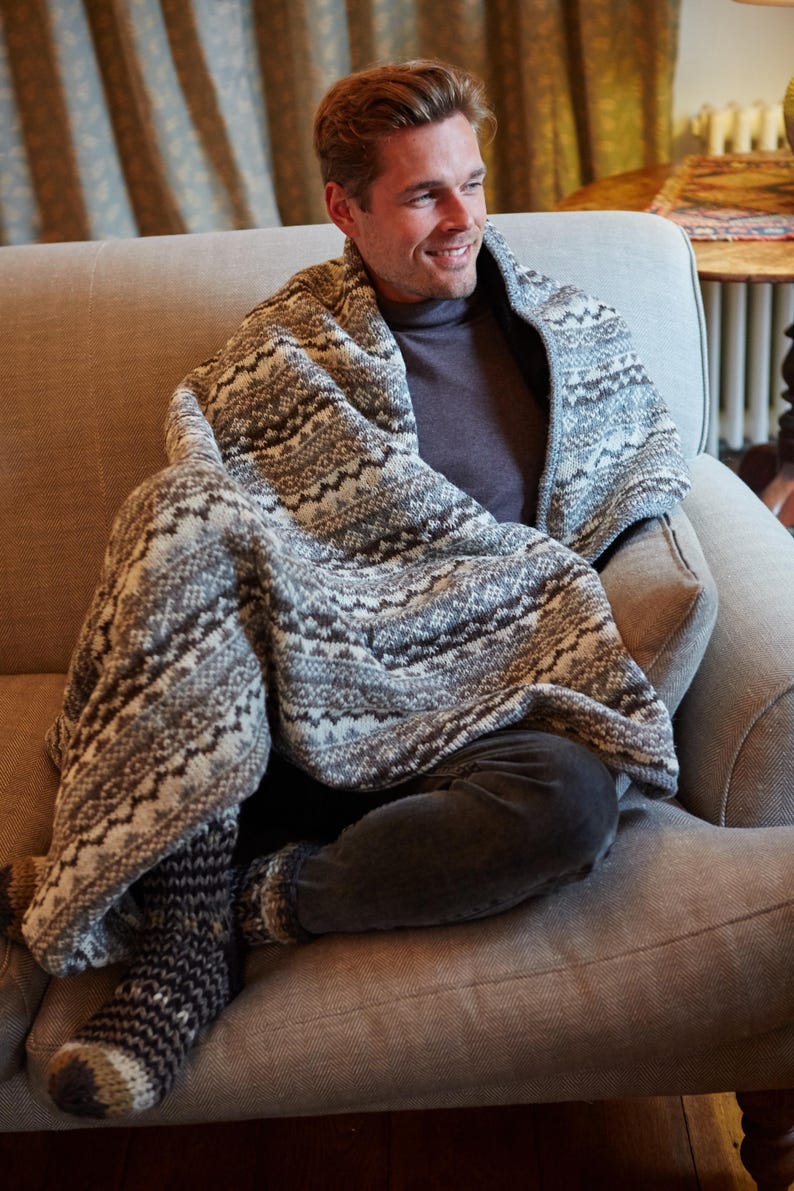 K&ouml;nnte beinhalten: Eine Person sitzt auf einem grauen Sofa und ist in eine Strickdecke mit grauem und wei&szlig;em Muster geh&uuml;llt. Die Person tr&auml;gt einen grauen Rollkragenpullover und eine dunkelgraue Hose. Passende Stricksocken sind sichtbar.