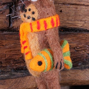 May include: A brown felt animal ornament with a red and orange scarf and a green and orange fish. The ornament is hanging from a wooden surface.