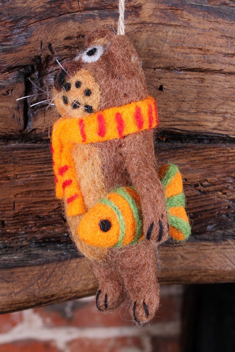 May include: A brown felt animal ornament with a red and orange scarf and a green and orange fish. The ornament is hanging from a wooden surface.
