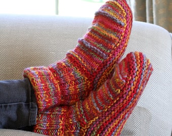 May include: A pair of colorful, hand-knitted slipper socks in shades of red, orange, yellow, and purple. The socks are thick and cozy, with a ribbed design on the upper part. They are resting on a gray herringbone patterned couch.