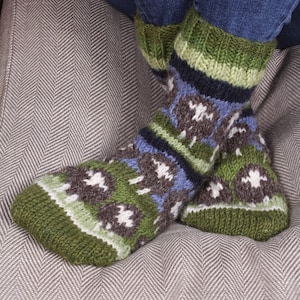 May include: A pair of hand-knitted wool socks featuring a repeating pattern of brown sheep with white woolly tops against a background of green, blue, and dark navy stripes. The socks have a ribbed cuff and a thick, textured knit construction.