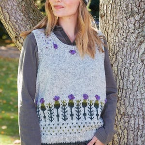 May include: A white knitted vest with a green and purple thistle pattern. The vest has a scoop neck and is sleeveless. The model is wearing a green and purple headband with a thistle pattern.