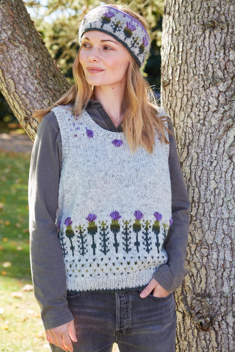 May include: A white knitted vest with a green and purple thistle pattern. The vest has a scoop neck and is sleeveless. The model is wearing a green and purple headband with a thistle pattern.