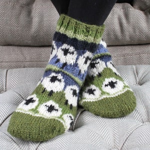 May include: Cozy, hand-knitted socks with a green, white, black, and blue pattern. The socks feature a repeating design of white shapes with black centers on a green background, with green cuffs and toes. The socks are worn on a gray cushioned surface.
