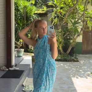 May include: A woman wearing a blue and white patterned sarong dress. She is standing in front of a brick wall and a green plant.