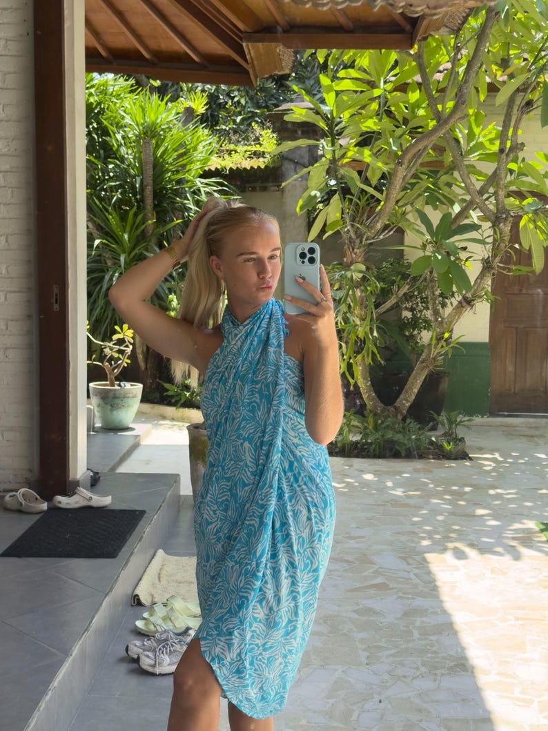 May include: A woman wearing a blue and white patterned sarong dress. She is standing in front of a brick wall and a green plant.