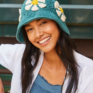 May include: A teal blue crochet hat with white and yellow daisy flowers. The hat has a wide brim and is worn by a person with long dark hair.