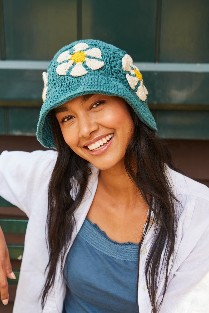 May include: A teal blue crochet hat with white and yellow daisy flowers. The hat has a wide brim and is worn by a person with long dark hair.