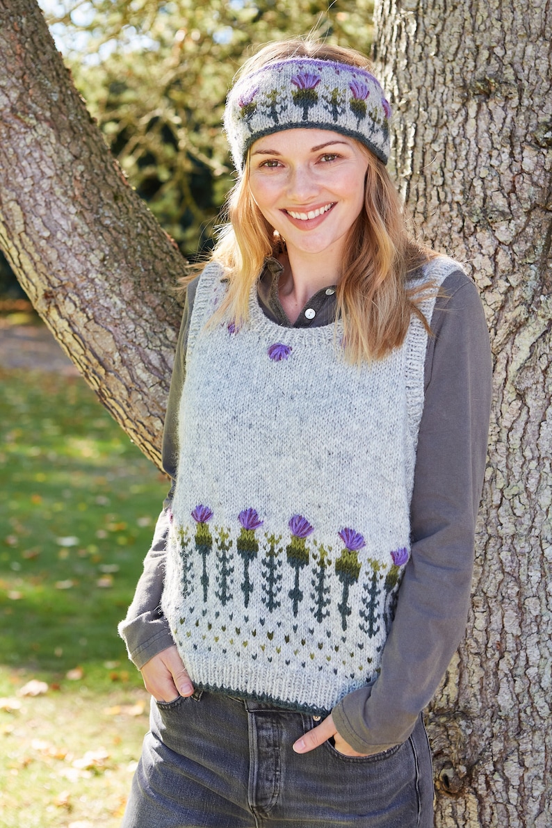 May include: A gray sleeveless sweater with a green and purple thistle pattern. The sweater has a round neckline and is worn over a gray long-sleeved shirt. The person is wearing a headband with a similar thistle pattern.