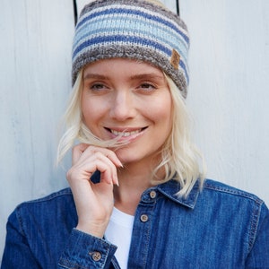 May include: A woman with blonde hair is wearing a blue and grey striped knit headband with a brown leather tag. She is smiling and wearing a blue denim shirt.