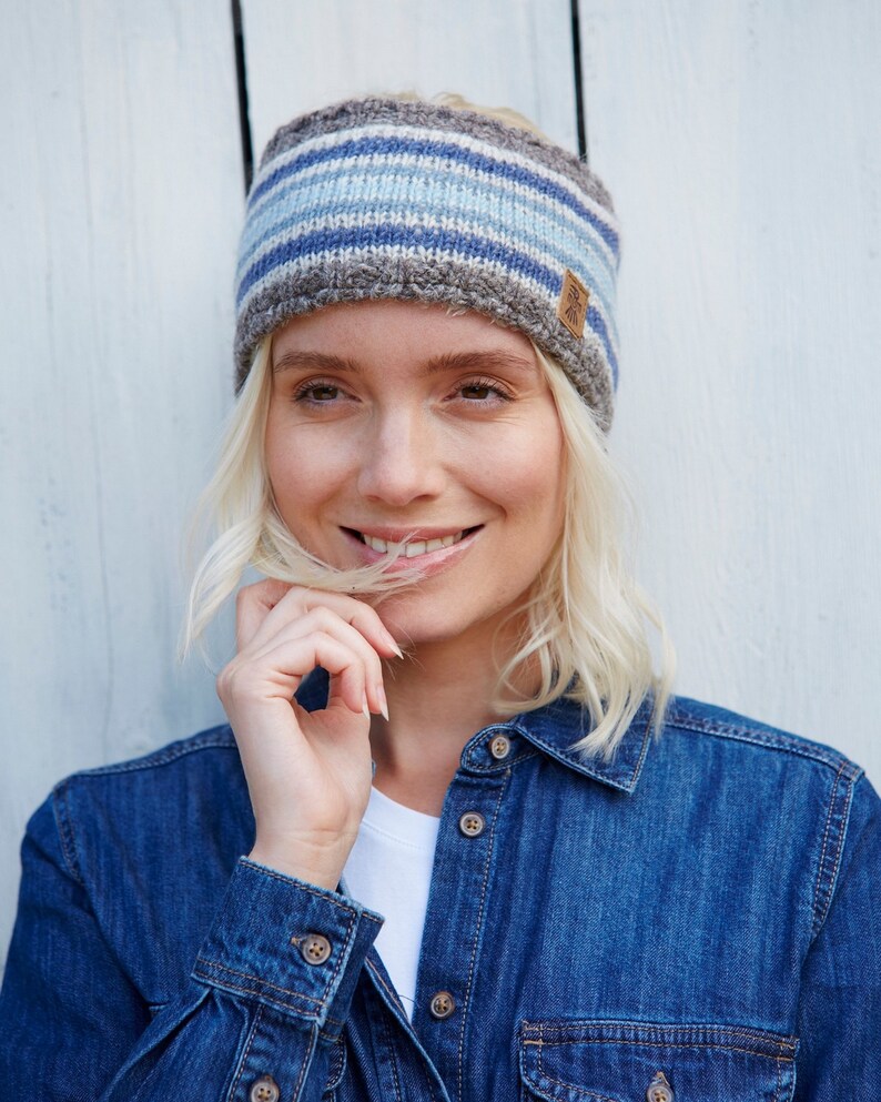 May include: A woman with blonde hair is wearing a blue and grey striped knit headband with a brown leather tag. She is smiling and wearing a blue denim shirt.