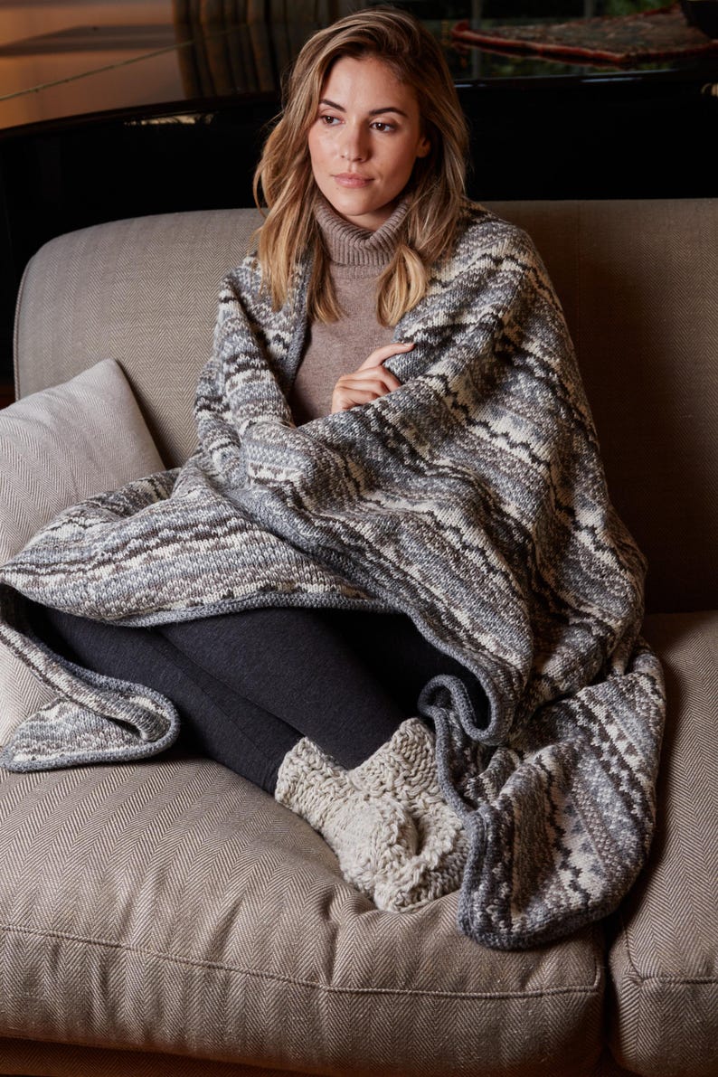 K&ouml;nnte beinhalten: Eine Person sitzt auf einem Sofa, eingeh&uuml;llt in eine grau-wei&szlig; gemusterte Decke. Die Decke hat ein geometrisches Design. Die Person tr&auml;gt graue Leggings und gestrickte Socken. Das Sofa hat eine neutrale Farbe.