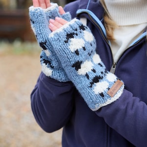 Könnte beinhalten: Hellblaue Strick-Fingerhandschuhe mit einem Schafmuster in Schwarz und Weiß. Die Handschuhe haben eine weiße Manschette und ein kleines braunes Lederetikett. Die Handschuhe sind aus einem weichen, warmen Material.