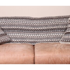 K&ouml;nnte beinhalten: Eine gestrickte Decke und zwei passende Kissen mit einem geometrischen Muster in Grau-, Braun- und Wei&szlig;t&ouml;nen werden auf einem braunen Ledersofa pr&auml;sentiert. Die Decke ist &uuml;ber die R&uuml;ckenlehne des Sofas drapiert, und die Kissen sind auf beiden Seiten platziert.