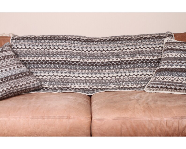 K&ouml;nnte beinhalten: Eine gestrickte Decke und zwei passende Kissen mit einem geometrischen Muster in Grau-, Braun- und Wei&szlig;t&ouml;nen werden auf einem braunen Ledersofa pr&auml;sentiert. Die Decke ist &uuml;ber die R&uuml;ckenlehne des Sofas drapiert, und die Kissen sind auf beiden Seiten platziert.