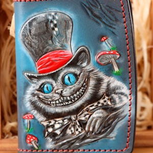 May include: A blue leather wallet with a grinning Cheshire Cat design, wearing a top hat with a red band. The wallet has red stitching and mushroom illustrations. The cat has blue eyes and a bow tie.