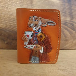 Puede incluir: Una cartera de cuero marrón claro con una ilustración detallada de un conejo vestido con un traje, sosteniendo una taza de té y un plato pequeño. El conejo tiene un girasol en la solapa y una corbata estampada. La cartera está cosida por los bordes.