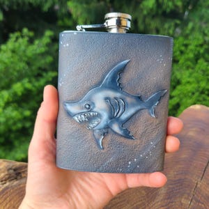 Puede incluir: Una petaca de metal rectangular con un acabado gris oscuro texturizado que presenta un diseño de tiburón en relieve en la parte frontal. El tiburón se muestra de perfil con dientes y aletas visibles. La petaca tiene un tapón de rosca plateado.