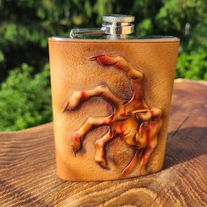 May include: A tan leather-wrapped metal flask featuring a three-dimensional embossed design of a clawed hand with red accents. The flask has a smooth, silver-coloured metal screw-top cap and a textured, light brown leather exterior surface.