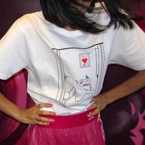 May include: A white t-shirt with a black and white illustration of a person in a doorway. The illustration includes the text "Self Love". The t-shirt is paired with a pink skirt.
