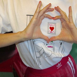 May include: A white t-shirt with a black and white illustration of a person in bed. The shirt has the words "Self Love" with a red heart in the center.