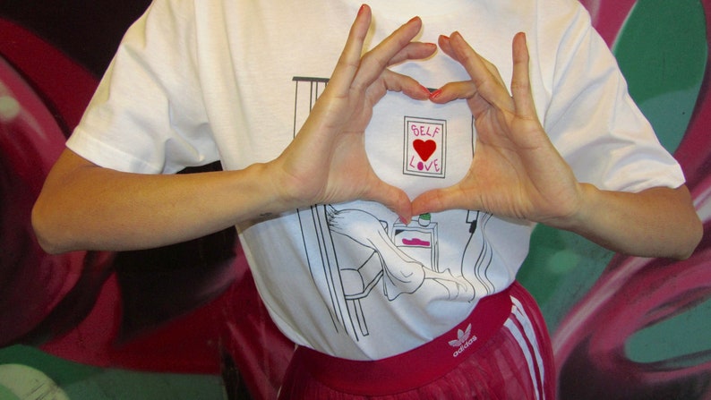 May include: A white t-shirt with a black and white illustration of a person in bed. The shirt has the words "Self Love" with a red heart in the center.