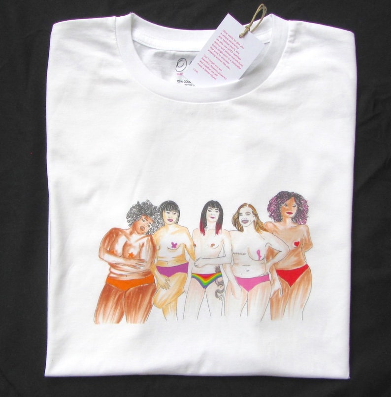 Puede incluir: Camiseta blanca con un gr&aacute;fico de cinco mujeres de pie juntas, cada una con un peinado y un tipo de cuerpo diferente. Las mujeres llevan ropa interior de varios colores y tienen cintas rosas en el pecho. El texto "100% Algod&oacute;n" est&aacute; impreso en la camiseta.