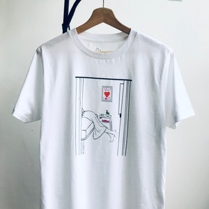 May include: White t-shirt with a black line drawing of a person in bed with a sign that says "Self Love" above the bed.