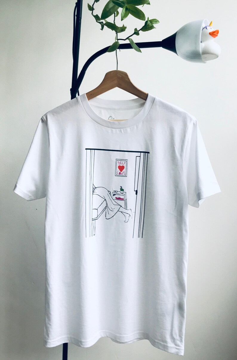 May include: White t-shirt with a black line drawing of a person in bed with a sign that says "Self Love" above the bed.