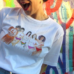 Puede incluir: Camiseta blanca con una ilustraci&oacute;n colorida de seis mujeres de pie juntas, cada una con los brazos alrededor de la otra. Las mujeres son de diferentes razas y tipos de cuerpo.