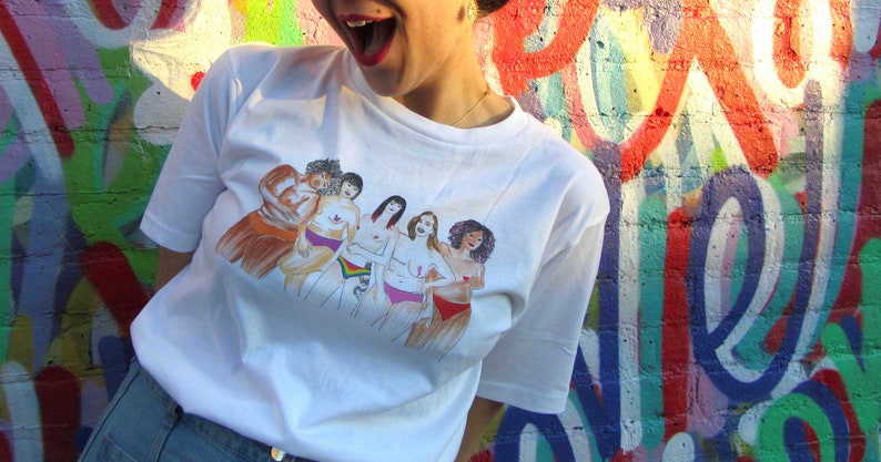 Puede incluir: Camiseta blanca con una ilustraci&oacute;n colorida de seis mujeres de pie juntas, cada una con los brazos alrededor de la otra. Las mujeres son de diferentes razas y tipos de cuerpo.