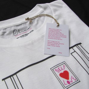 May include: White t-shirt with a black and white illustration of a picture frame with the words "SELF LOVE" inside. The heart in the frame is red. The t-shirt has a tag attached with the text "Olina wants to give you a unique experience with fashion that's why we only use GOTS certified fabrics and ethical manufacture. Every single print is a storyteller that aims to empower amazing women like you. Don't forget you are beautiful, flawless, powerful and together we will make a better future! Love, Olina"