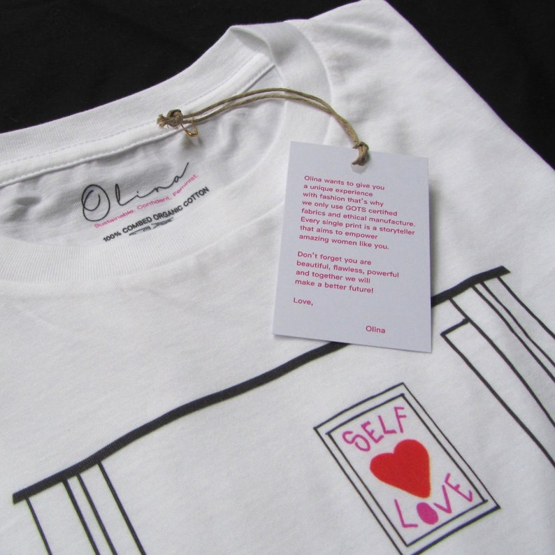 May include: White t-shirt with a black and white illustration of a picture frame with the words "SELF LOVE" inside. The heart in the frame is red. The t-shirt has a tag attached with the text "Olina wants to give you a unique experience with fashion that's why we only use GOTS certified fabrics and ethical manufacture. Every single print is a storyteller that aims to empower amazing women like you. Don't forget you are beautiful, flawless, powerful and together we will make a better future! Love, Olina"