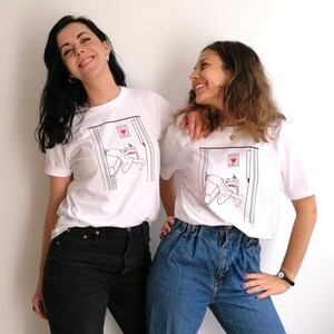 May include: Two people wearing white t-shirts with a graphic of a person sleeping in a bed with a heart on the door. The person on the left is wearing black jeans and the person on the right is wearing blue jeans.
